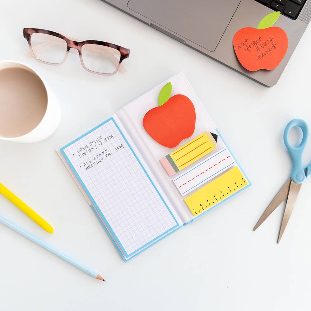 Open note set on a desk beside glasses, a coffee mug, scissors and a laptop with an apple-shaped note stuck on it
