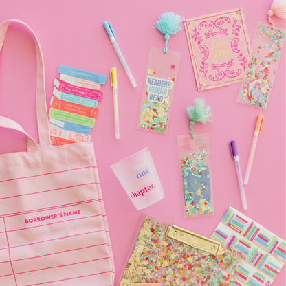 Confetti bookmarks with pink and aqua tassels on a pink table beside a tote bag, pens and a clutch