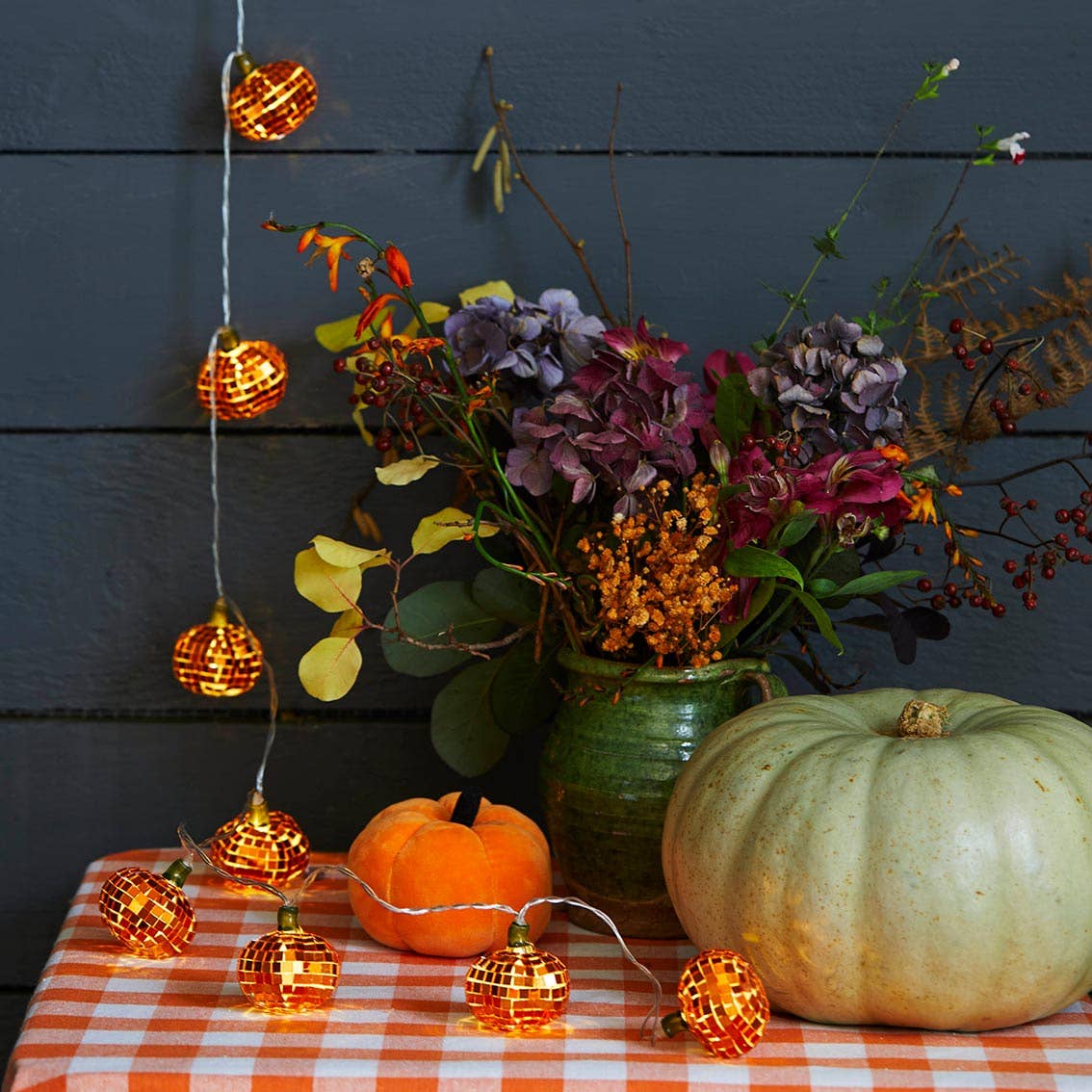 Orange mirror-ball pumpkin lights hanging beside a vase of flowers and pumpkins on a checked tablecloth
