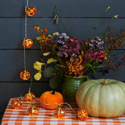 Orange mirror-ball pumpkin lights hanging beside a vase of flowers and pumpkins on a checked tablecloth