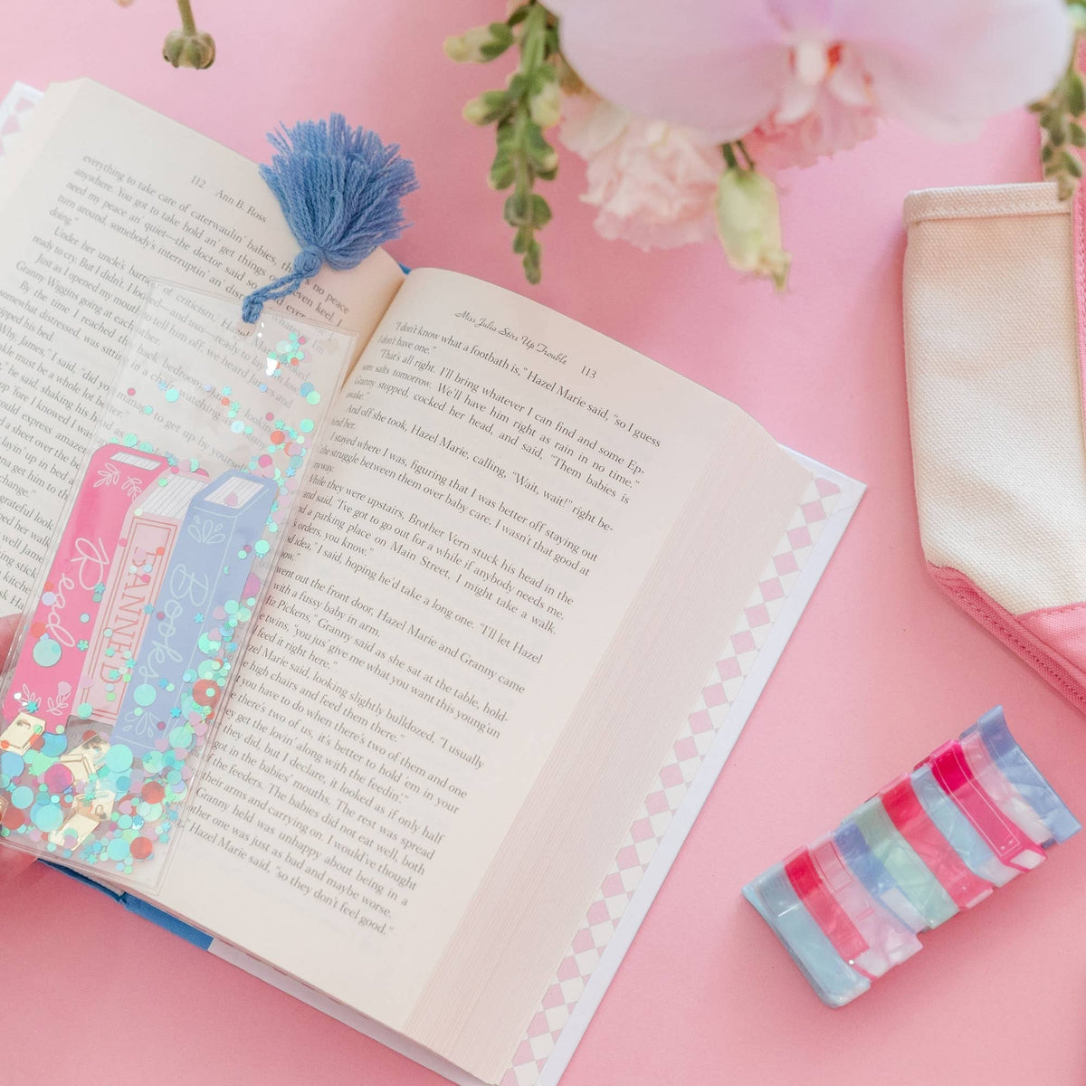 Confetti bookmark with a blue tassel resting on an open book beside pink flowers