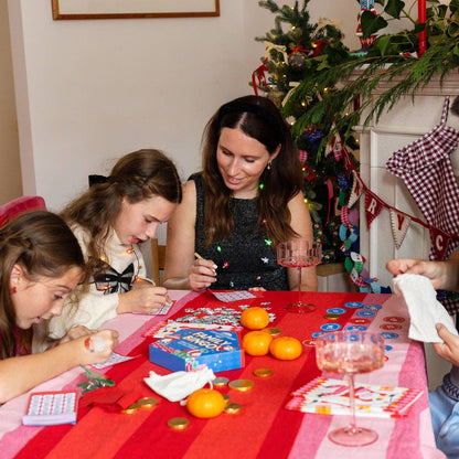 Family playing bingo at a red table with oranges and a pink glass near a Christmas tree