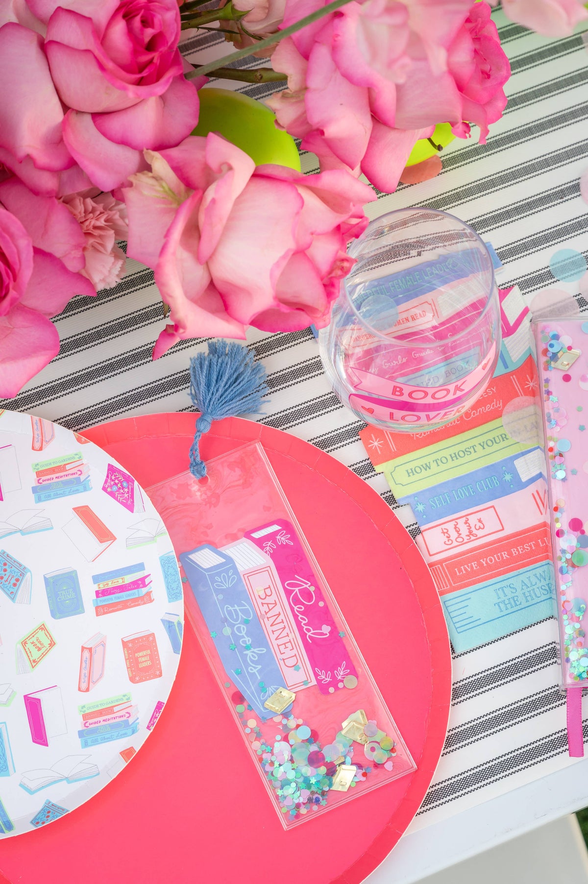 Confetti bookmark with a blue tassel on a striped tablecloth beside roses, a glass and a patterned plate
