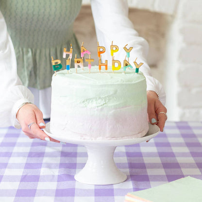 Cake with rainbow Happy Birthday letter candles being carried to a table with a purple gingham cloth