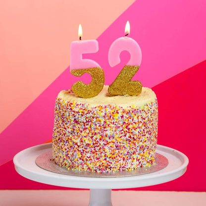 Pink and gold glitter number candles reading 52, lit on a sprinkle-covered cake against a pink backdrop